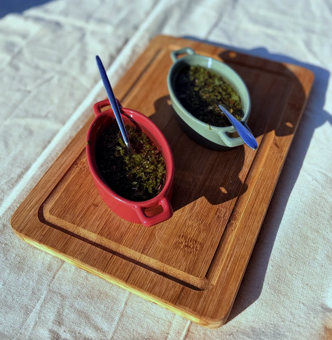 Two bowls of chimichurri on a wooden board, spoons in